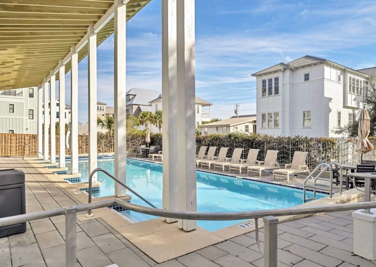 A pool area with a covered deck, lounge chairs, and a view of surrounding buildings.