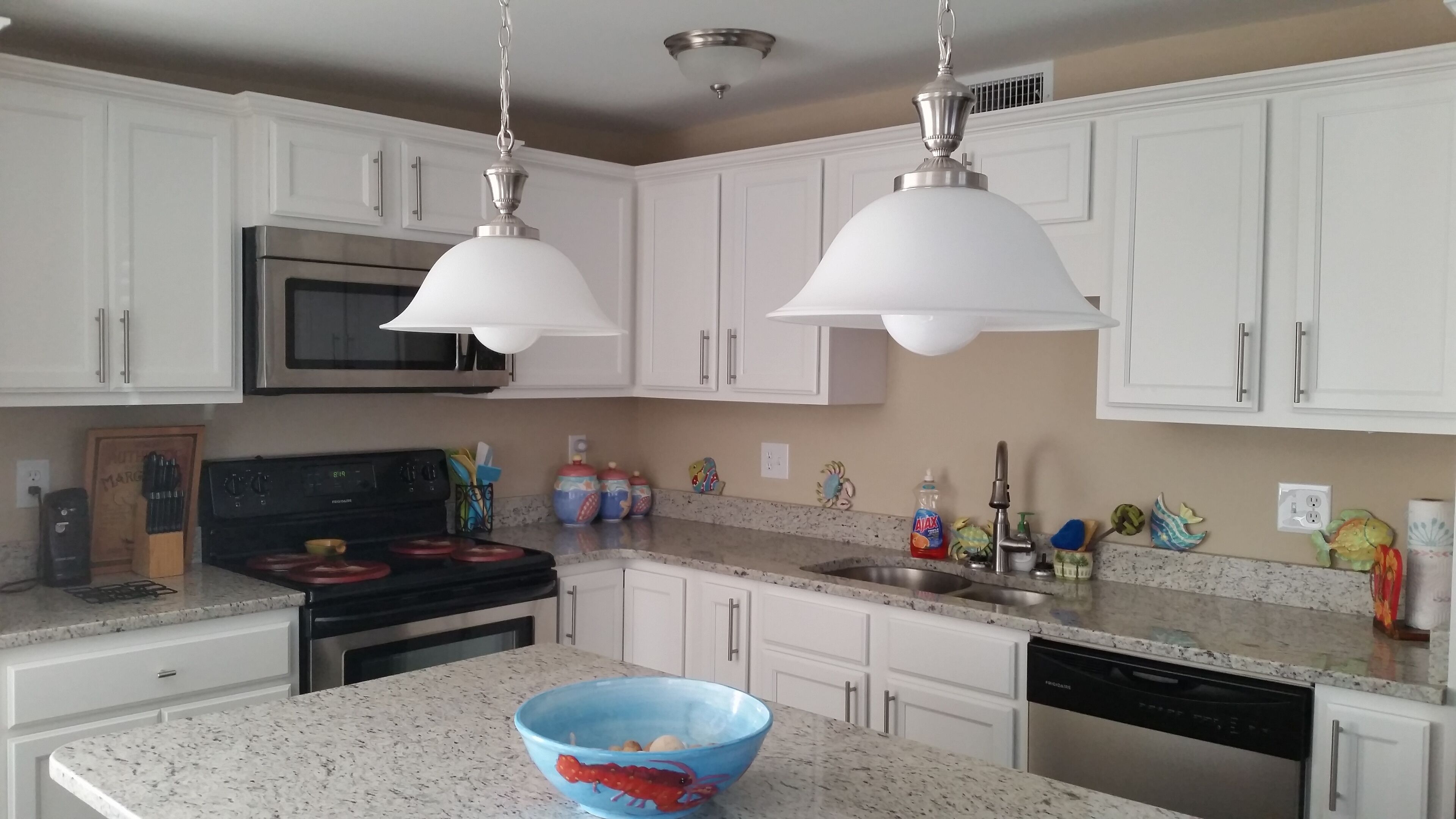 A kitchen with white cabinets, a stainless steel stove, a microwave, and a dishwasher. There are two pendant lights, a blue bowl on the counter, and various kitchen utensils and decorations.
