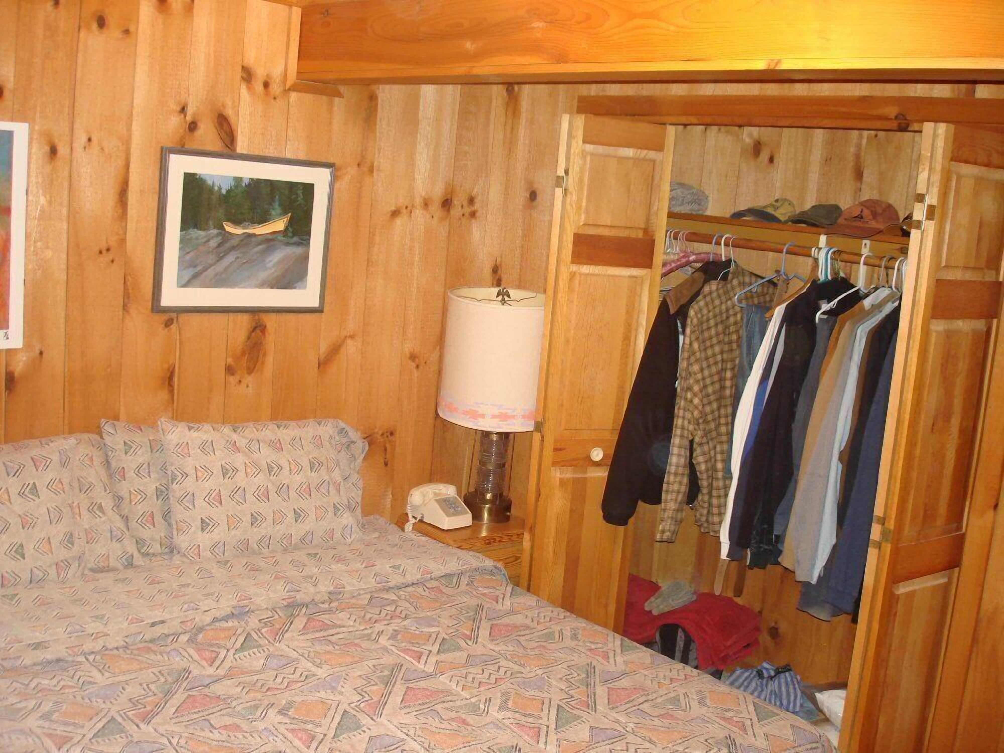 Una habitación de madera con una cama, una lámpara, un perchero y una puerta que da a otra habitación.