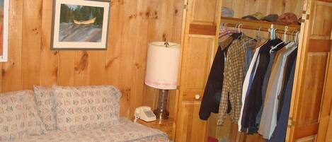 Una habitación de madera con una cama, una lámpara, un perchero y una puerta que da a otra habitación.