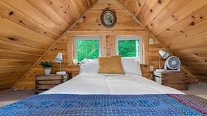 Quarto de sótão acolhedor com teto inclinado de madeira, uma cama grande com roupa de cama branca, duas janelas, uma mesinha com um abajur e um ventilador.