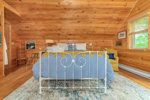 A cozy cabin-style room with a bed, wooden walls, a window, and a small table with a lamp.