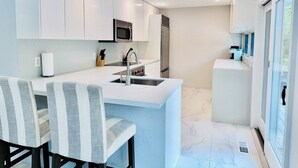 A modern kitchen with white cabinetry, a central island with seating, stainless steel appliances, and a large window providing natural light.