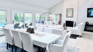 A spacious living room with a dining area, white furniture, a fireplace, and large windows overlooking a deck.