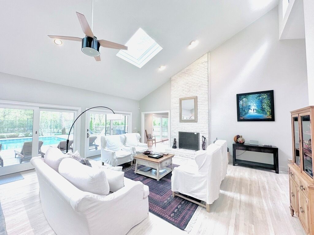 A spacious living room with white furniture, a ceiling fan, a fireplace, and a large window overlooking a pool.