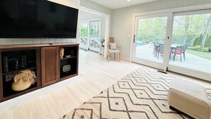 A spacious living room with a large flat-screen TV, wooden cabinets, a patterned rug, and a sliding glass door leading to an outdoor area.