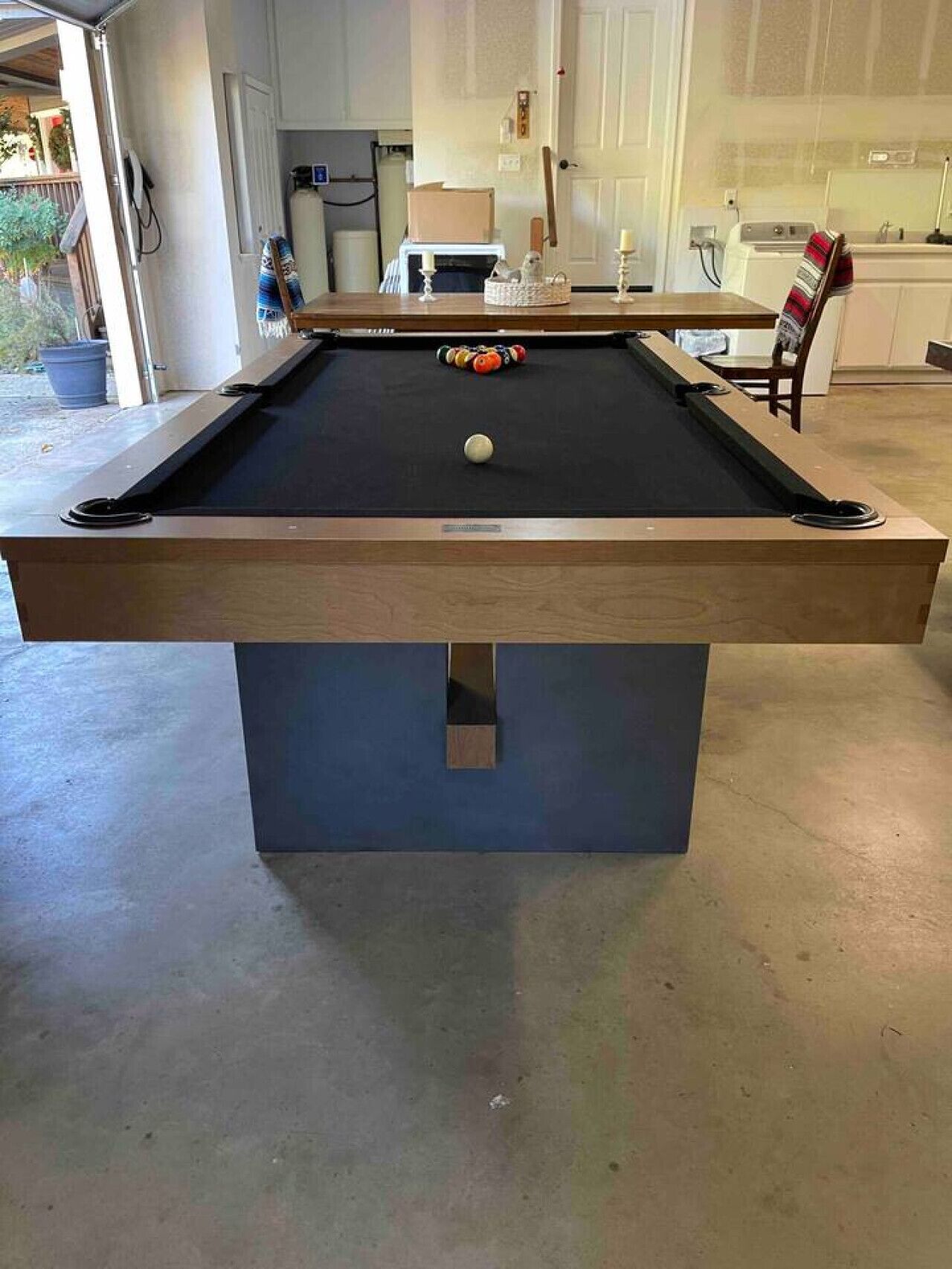 A pool table in a room with a concrete floor, a door, and a kitchen area in the background.
