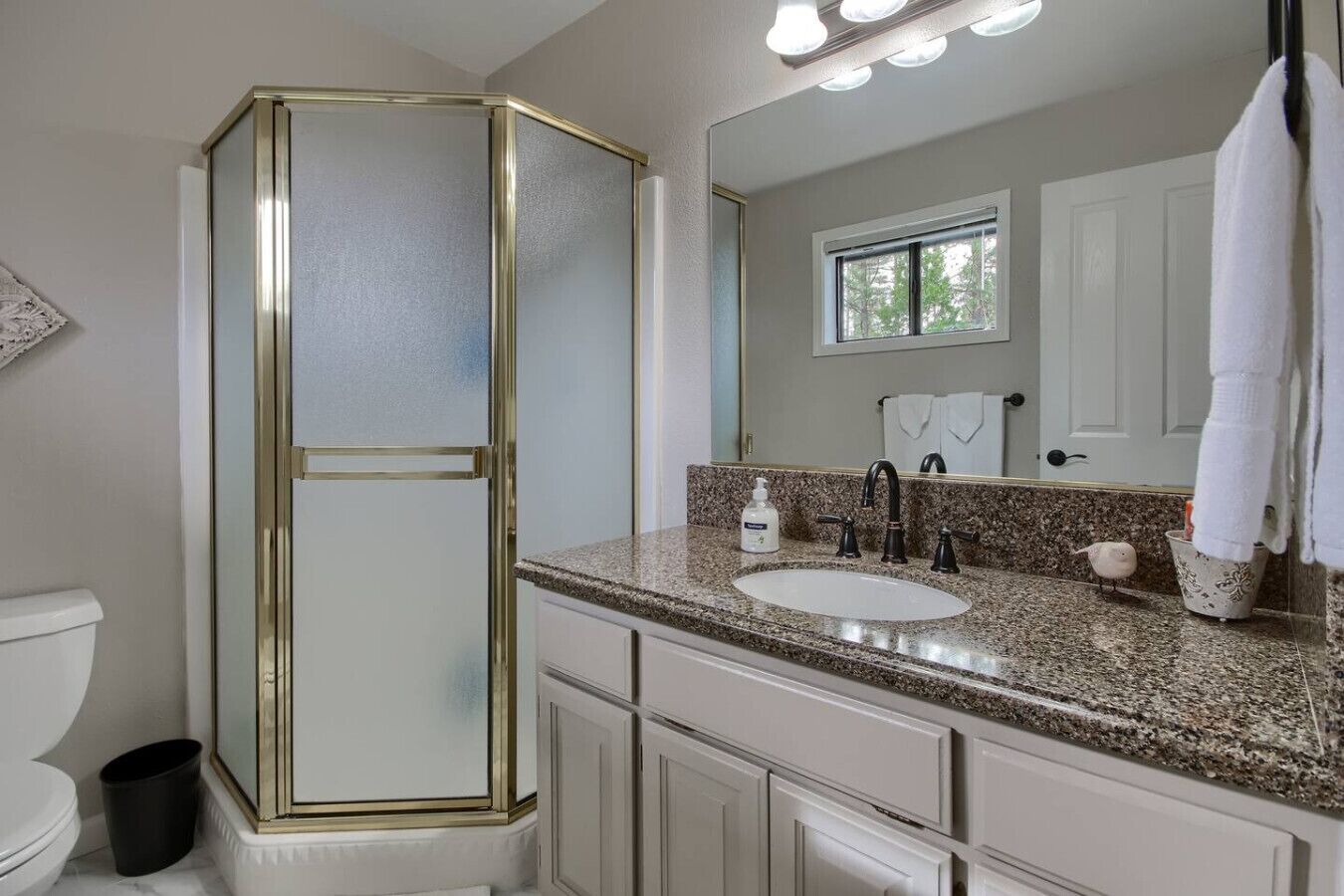 Une salle de bain avec des toilettes blanches, un lavabo doté d’un plan de travail en granit, un miroir et une douche avec un cadre doré.