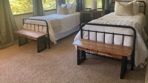 A bedroom with two single beds, a wooden bench, a bedside table, a lamp, and a TV on the wall.