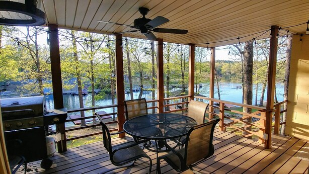 A covered outdoor deck with a table and chairs, overlooking a body of water and trees.