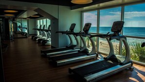 Una palestra con tapis roulant e attrezzature per esercizi, ampie finestre vista spiaggia.
