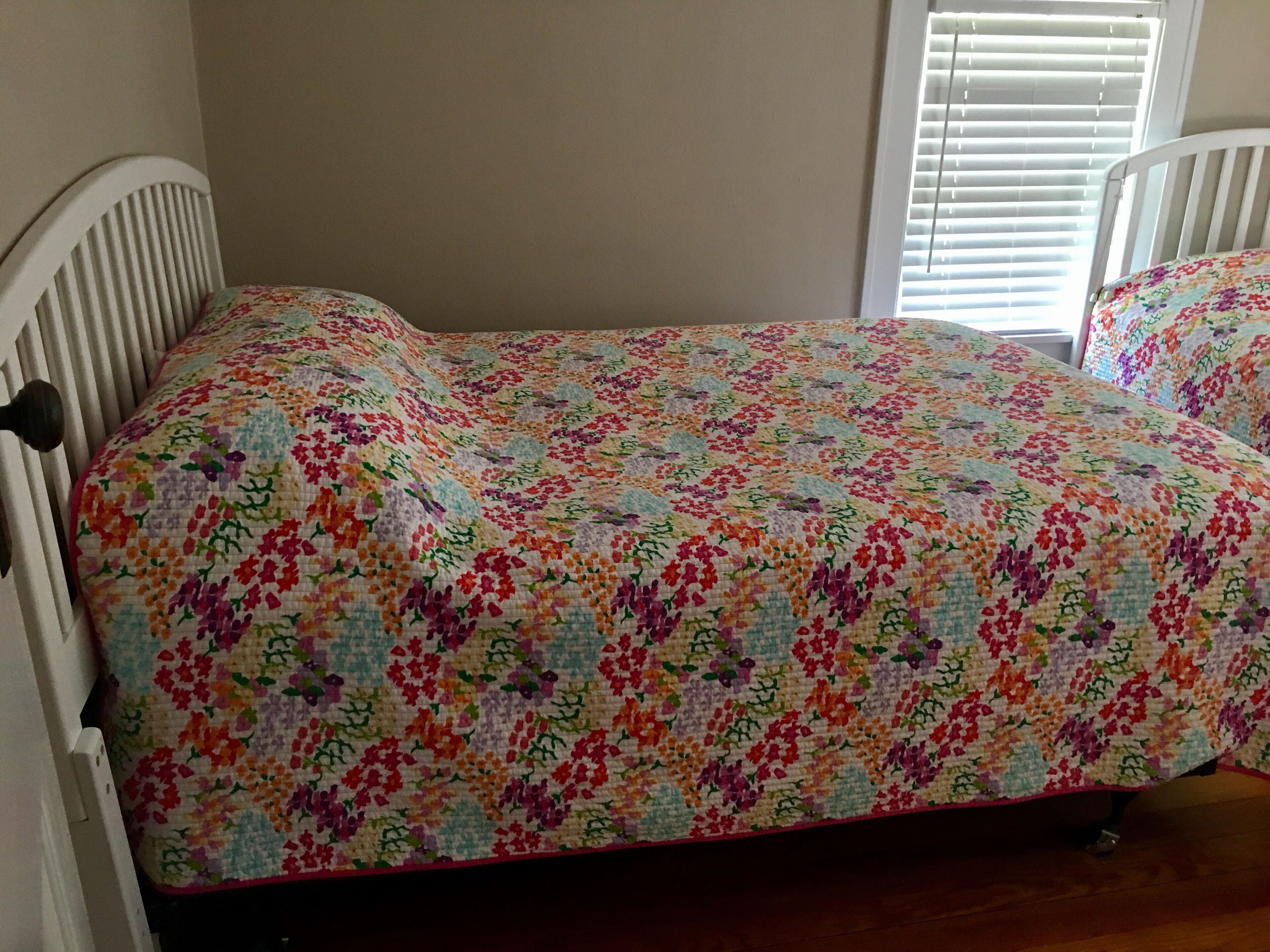Ein Bett mit floraler Steppdecke, weißem Kopfteil und einem Fenster mit Jalousien.