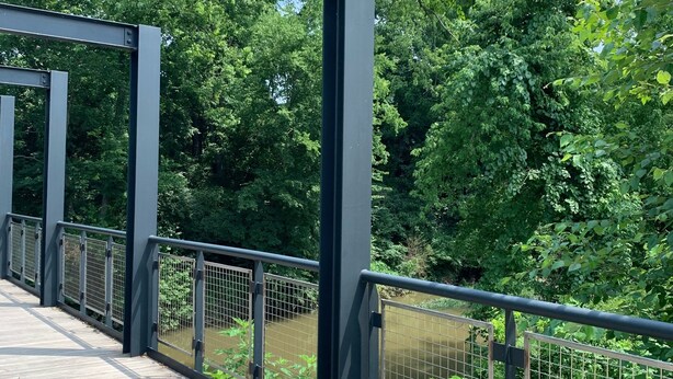 A wooden pedestrian bridge with metal railings, surrounded by lush greenery and trees.