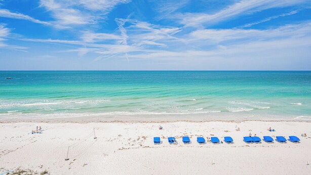 A beach with white sand and a clear blue sky. There are blue lounge chairs lined up on the sand.