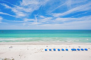 Een strand met wit zand en een helderblauwe lucht. Er staan blauwe ligbedden op een rij in het zand.