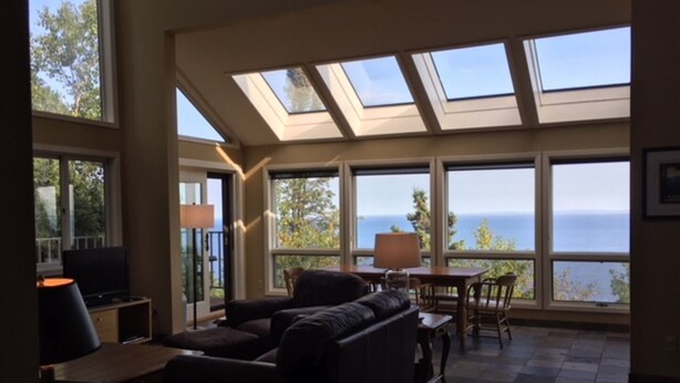 A spacious living room with a large sofa, a television, and a table with chairs. There are large windows offering a view of the ocean. - Unparalleled 180-degree Views of Lake Superior, 400 feet above the water! (Two Harbors)
