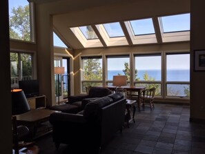 A spacious living room with a large sofa, a television, and a table with chairs. There are large windows offering a view of the ocean.
