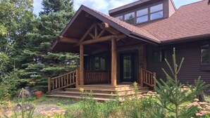 A wooden cabin with a porch, surrounded by dense forest and flowering plants. - Unparalleled 180-degree Views of Lake Superior, 400 feet above the water! (Two Harbors)