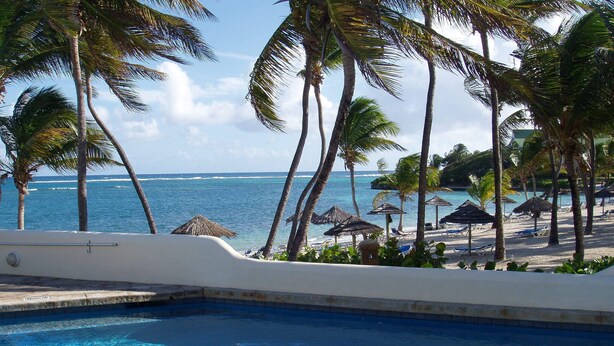 Una piscina con agua azul cristalina, rodeada de palmeras y sombrillas de playa, con vista a una playa y al océano.