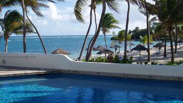 A pool with clear blue water, surrounded by palm trees and beach umbrellas, overlooking a beach and ocean.