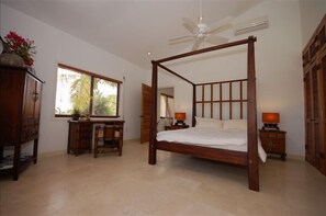 A bedroom with a four-poster bed, a desk with a chair, a window with a view of palm trees, and a ceiling fan.