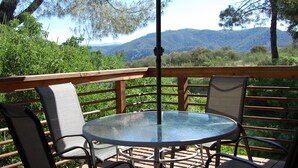 A round glass table with metal chairs and a wooden railing, overlooking a mountainous landscape.