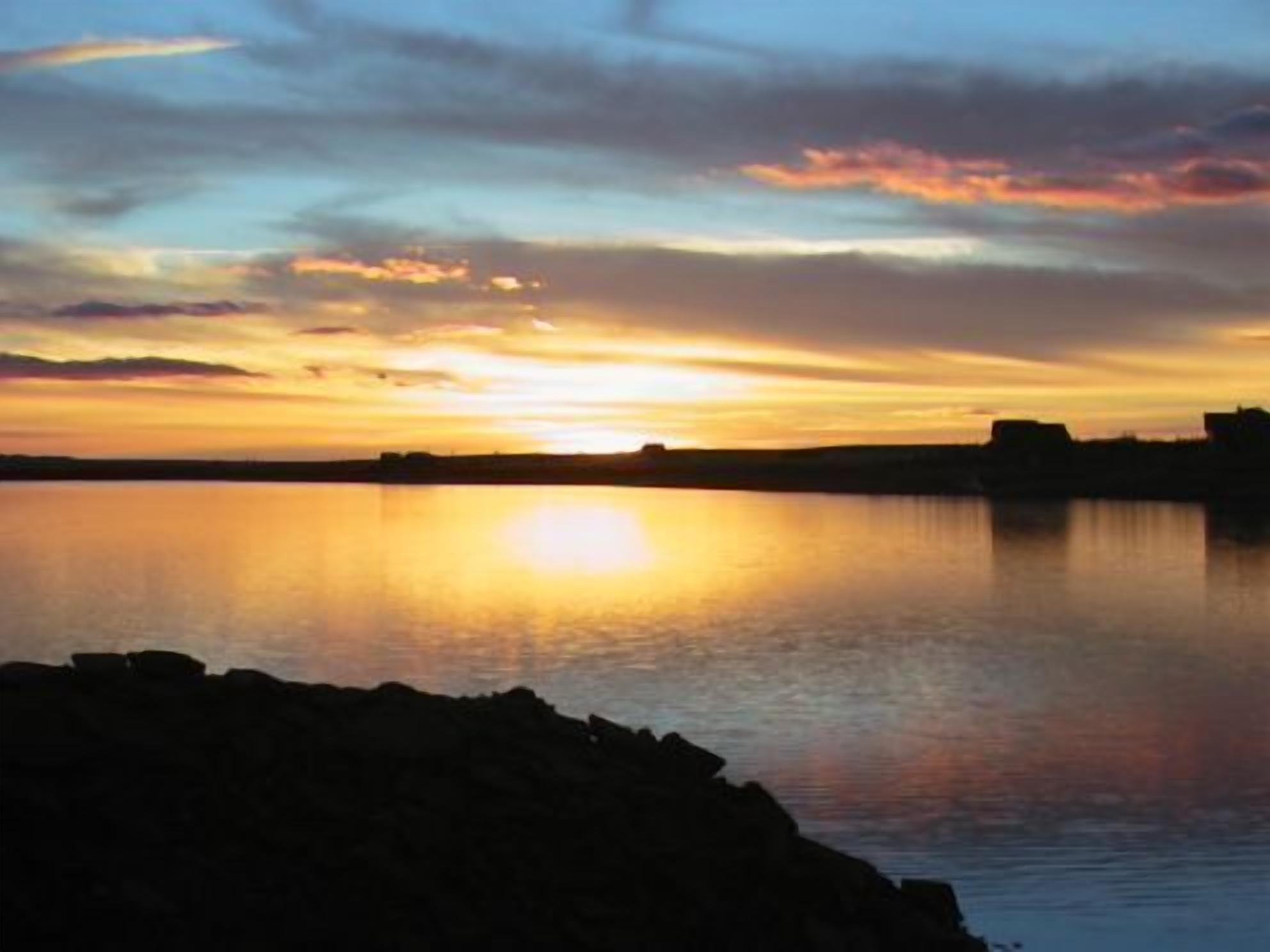 Un coucher de soleil sur une étendue d’eau calme, avec en silhouette une côte rocheuse et des bâtiments éloignés.