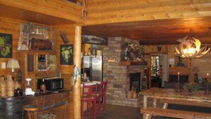 Interior rústico de una cabaña con chimenea de piedra, paredes de madera, una mesa de comedor y una zona de cocina con refrigerador y taburetes de bar.