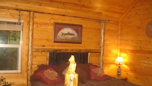 Habitación de cabaña de madera con una cama, un cuadro enmarcado de un pez, una ventana y una mesita de noche con una lámpara.