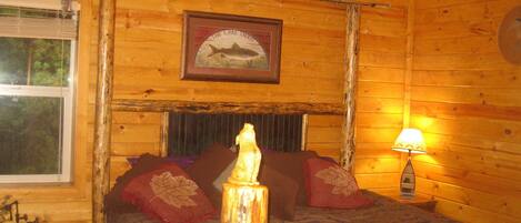 Habitación de cabaña de madera con una cama, un cuadro enmarcado de un pez, una ventana y una mesita de noche con una lámpara.
