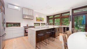 A modern kitchen with a central island, bar stools, and large windows offering a view of greenery. - West Coast Designer Home - Ocean & Forest (District of North Vancouver)