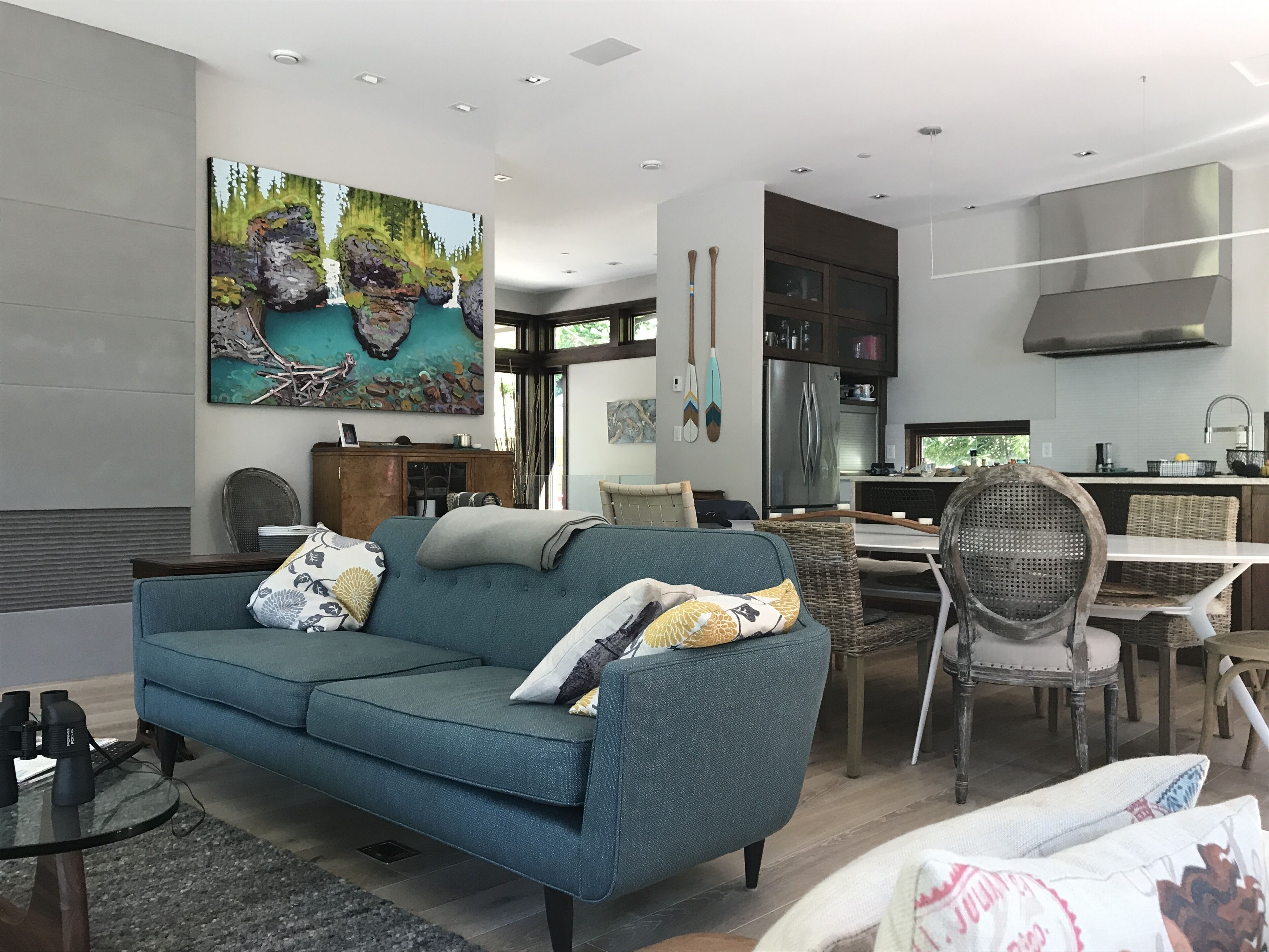 A modern living room with a blue sofa, a dining area with wicker chairs, a kitchen with stainless steel appliances, and a large painting on the wall.