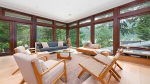 A spacious living room with a large window, a sofa, a coffee table, and a bookshelf. - West Coast Designer Home - Ocean & Forest (District of North Vancouver)