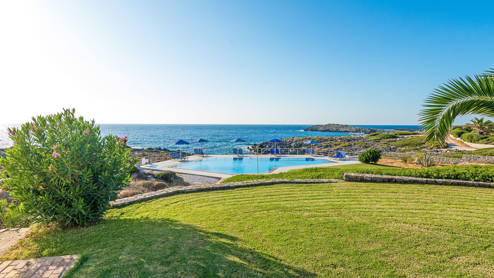 A pool area with a view of the sea, surrounded by greenery and a stone wall.