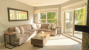 A living room with a beige sectional sofa, a wooden coffee table, and a large window with a view of greenery.
