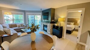 A modern living room with a stone fireplace, a dining area with a round table, and a bedroom with a large bed and headboard.