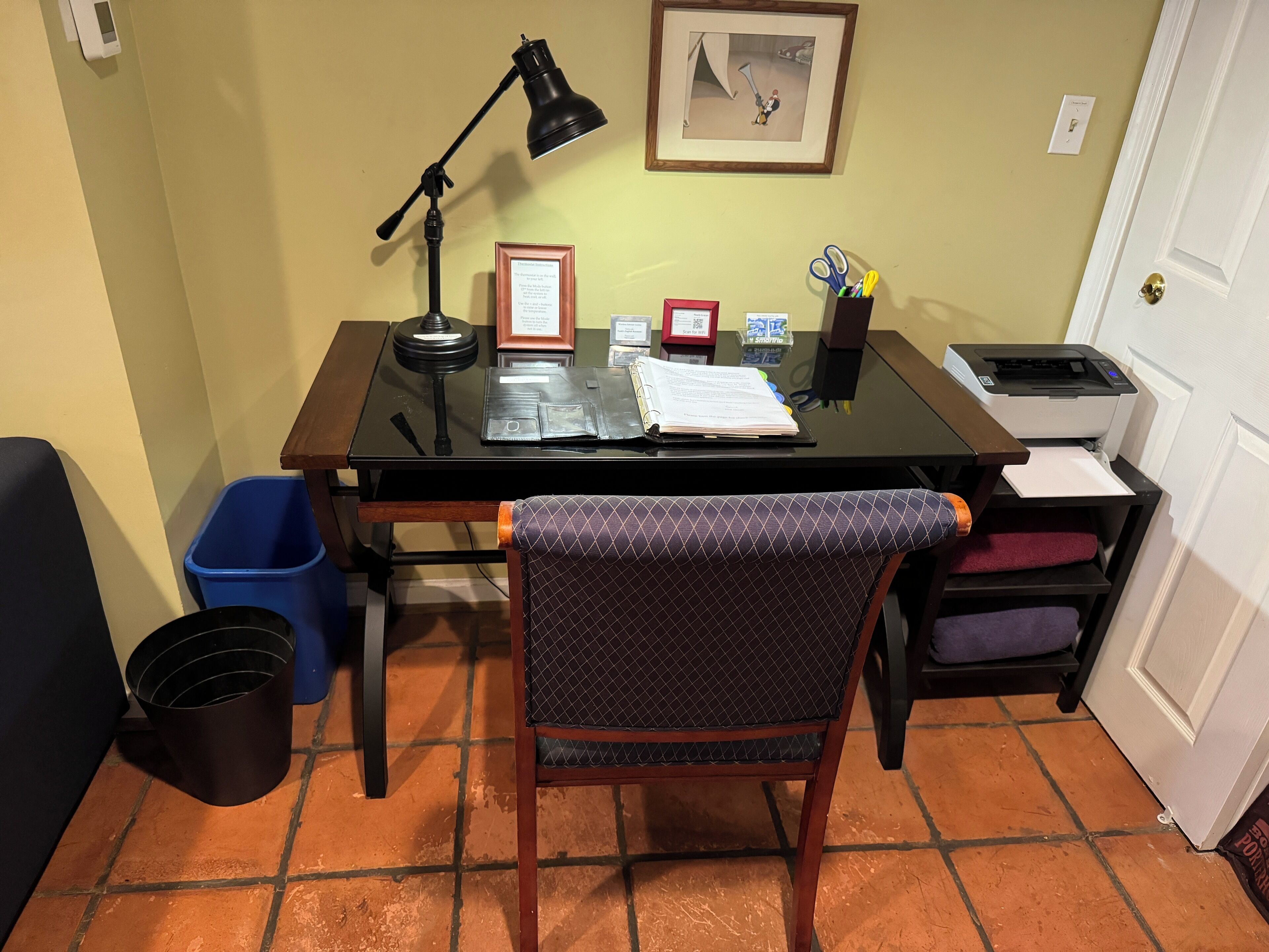Un petit bureau comprenant un bureau, une chaise, une lampe, des certificats encadrés et une imprimante.