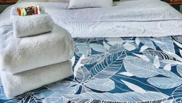 Un lit recouvert d’une couvre-lit blanche et bleue à motifs de feuilles, un oreiller orné du texte « BEACH LIFE », et une table de chevet avec une lampe et un objet décoratif.