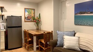 Une cuisine compacte comprenant un réfrigérateur, une table à manger en bois et des chaises. Un tableau représentant un paysage tropical orne le mur, ainsi qu’une affiche illustrant une plage.