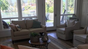 Ett vardagsrum med en beige soffa, ett soffbord med dekorativa föremål och utsikt över en sjö och hus genom stora fönster.