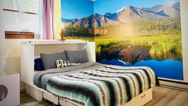 Ein Schlafzimmer mit Bett, einem Fenster mit Vorhängen und einer großen Wandmalerei von einer Berglandschaft.
