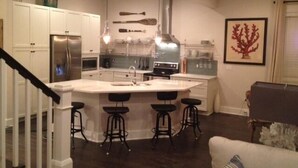 A modern kitchen with white cabinets, a stainless steel refrigerator, a large island with bar stools, and a hanging light fixture. - Waterfront Home with Golf Cart, Kayaks, Bikes and Paddleboards! (Navarre Beach)