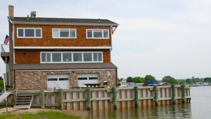 A two-story house with a stone facade, a dock extending into a body of water, and a flag on the left side. - Oyster lovers wanted! One of a kind property with 180 degree waterviews! (Mystic)