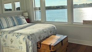A bedroom with a large bed, a wooden chest, and a view of a lake through large windows. - Oyster lovers wanted! One of a kind property with 180 degree waterviews! (Mystic)