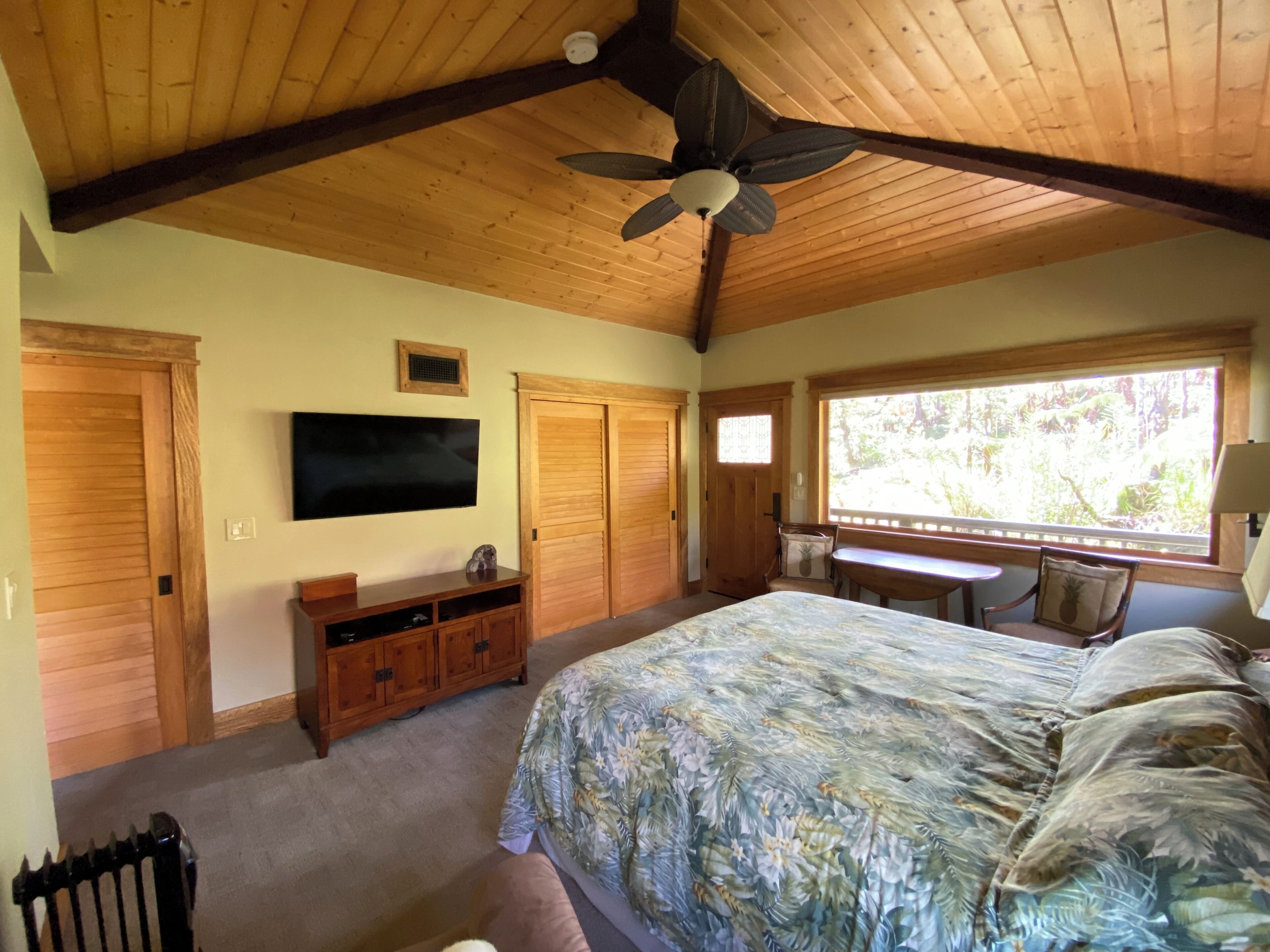 Ein Schlafzimmer mit einem großen Bett, einem Holzschrank, einem an der Wand montierten Fernseher und einem Fenster mit Blick auf Grünflächen.