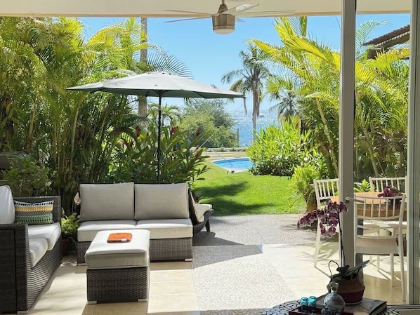 Une terrasse ensoleillée, dotée d’un canapé, de chaises et d’un parasol, qui offre une vue sur une piscine et une végétation luxuriante.
