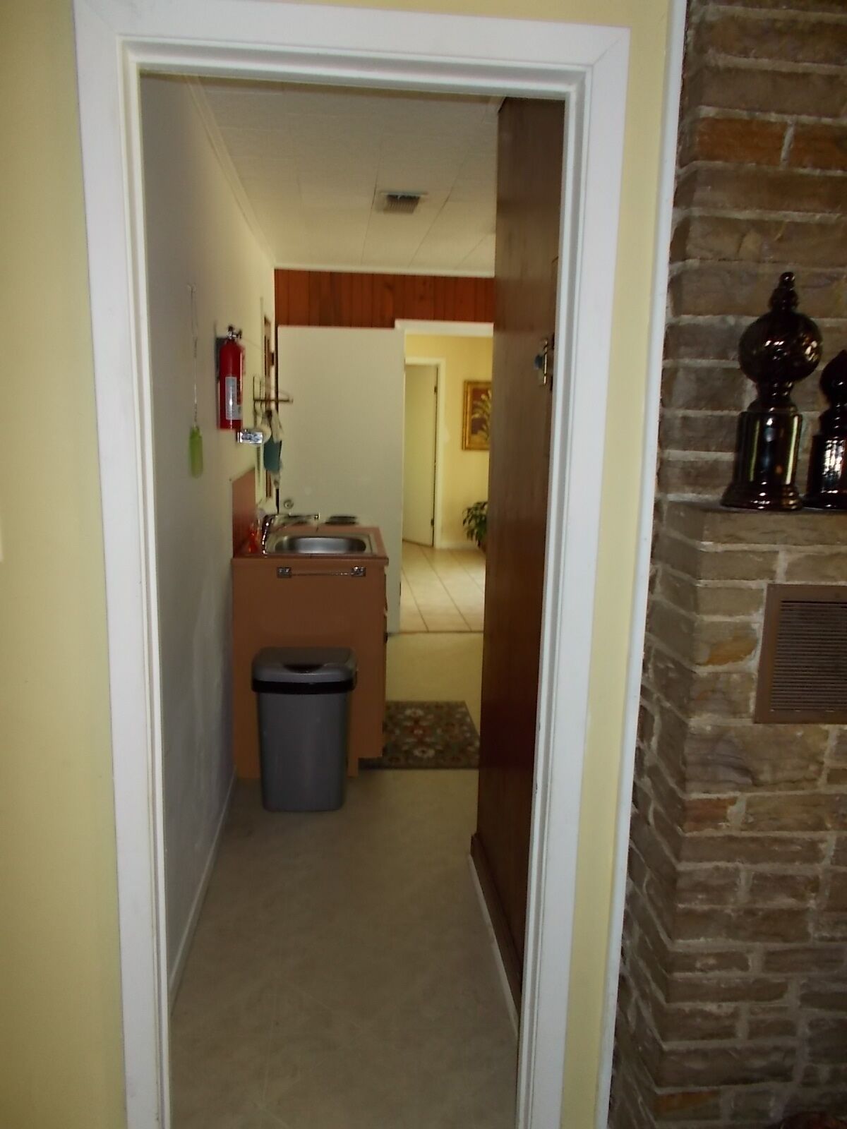 Un couloir étroit, avec une petite cuisine au fond, une poubelle et une cheminée agrémentée d’objets décoratifs.