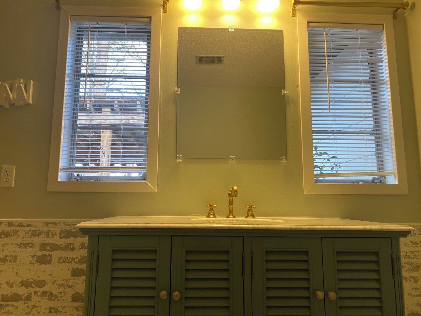 A bathroom with a sink, two faucets, and a mirror above two windows with blinds.