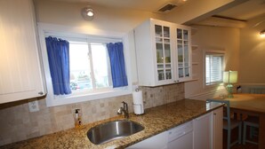 Una cocina con encimera de granito, pileta de acero inoxidable, muebles blancos y una ventana con cortinas azules.