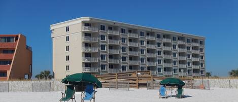 Hotel frente a la playa con varios balcones, una playa arenosa y sillas de playa bajo sombrillas.